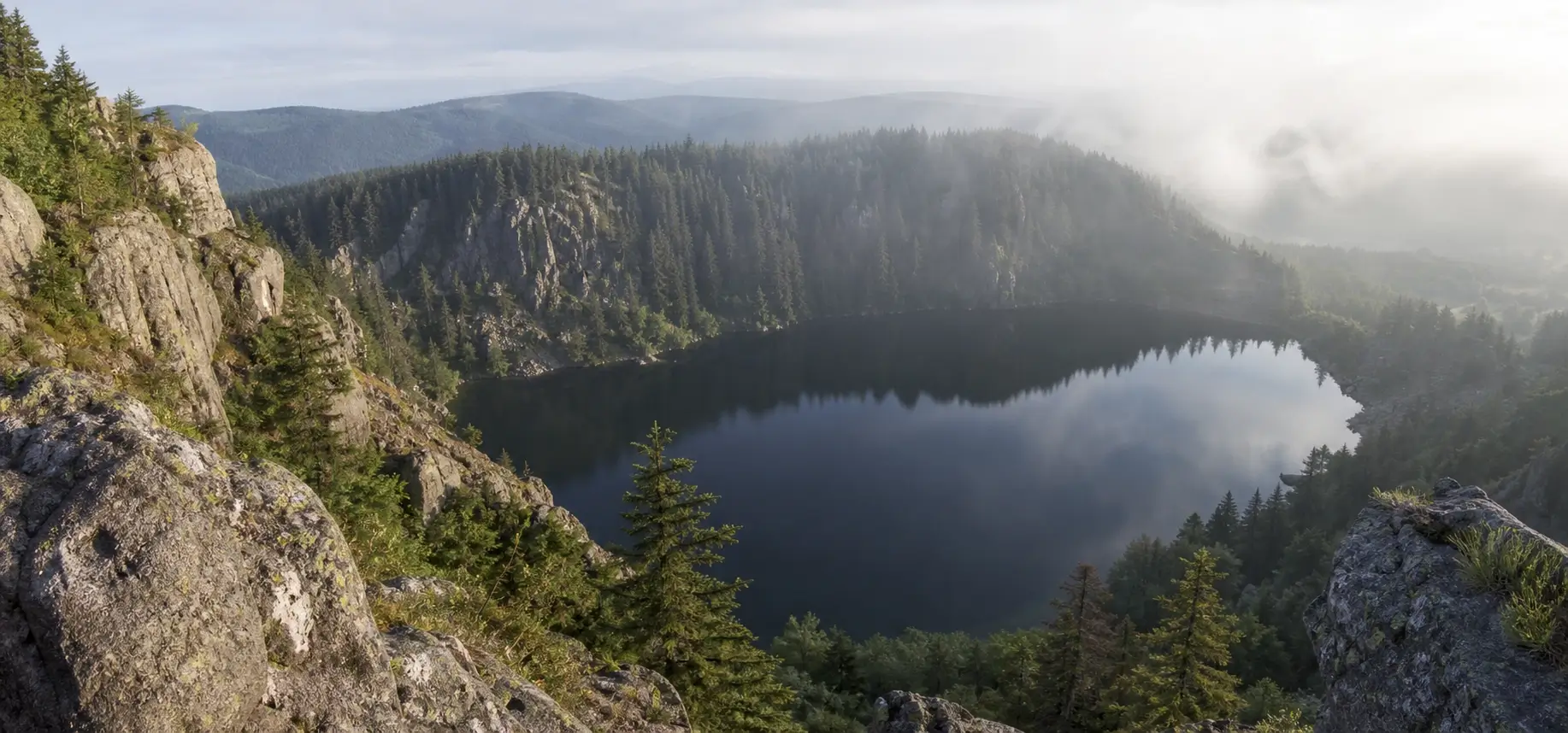 Vue du Lac Noir dans les Vosges entouré de falaises rocheuses et de forêt, avec une eau sombre et calme sous une lumière douce